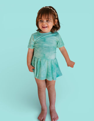 Toddler wearing jade tie-dye short-sleeve dress with gathered skirt against light blue background.