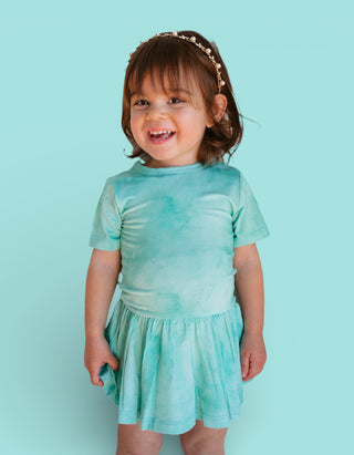 Toddler wearing jade tie-dye short-sleeve bodysuit dress with gathered skirt against turquoise background.