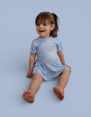Toddler wearing light blue tie-dye short-sleeve bodysuit dress with gathered skirt against blue background.