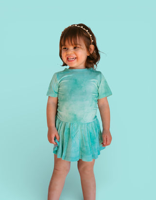 Toddler wearing a jade tie-dye short-sleeve dress with gathered skirt against a light blue background.