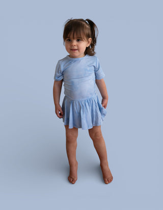 Toddler wearing light blue short-sleeve bodysuit dress with gathered skirt against blue background.