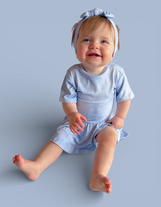 Toddler wearing light blue short-sleeve bodysuit dress with matching headbow, seated against blue background.