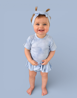 Toddler wearing light blue tie-dye short-sleeve bodysuit dress with matching bow headband against blue background.