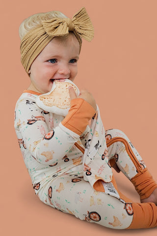 Infant holding cream and orange plush lovey with farm animal prints, wearing matching pajamas with tan bow headband.