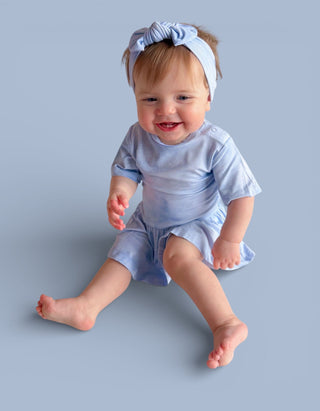 Smiling baby wearing light blue short-sleeve bodysuit dress with matching bow headband.