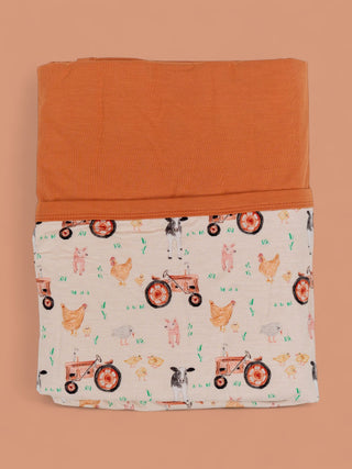 Folded blanket with orange solid top half and cream bottom half printed with tractors, chickens, pigs, and grass.