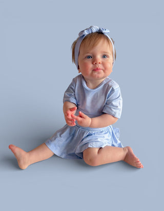 Infant wearing light blue short-sleeve bodysuit dress with matching headband, sitting against blue background.