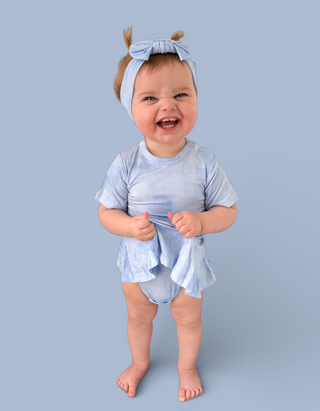 Toddler wearing light blue short-sleeve bodysuit dress with matching bow headband against blue background.