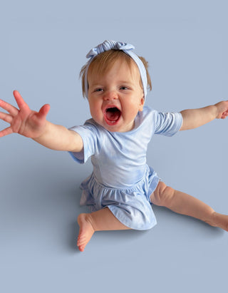 Toddler wearing light blue short-sleeve bodysuit dress with matching headband, arms outstretched against blue background.
